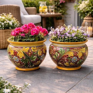Op de afbeelding: Twee decoratieve keramische bloempotten met kleurrijke bloemmotieven. De potten zijn gevuld met levendige roze en paarse bloemen. De potten hebben een gele rand en basis en staan op een terras.