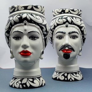 Puede incluir: Dos jarrones de cerámica blanca con diseños en blanco y negro. Los jarrones tienen forma de cabeza humana con rasgos estilizados. Un jarrón tiene una cara femenina con labios rojos y el otro tiene una cara masculina con barba y bigote negros.