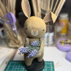 Puede incluir: Una figura de conejo artesanal de color beige, con orejas largas y caídas y una cara cosida. El conejo sostiene un pequeño ramo de flores de lavanda. Lleva un peto y se apoya sobre una base negra. El fondo está borroso, mostrando materiales de arte.