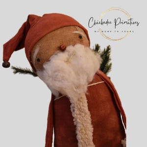 Può includere: Una bambola artigianale di Babbo Natale con un cappotto e un cappello color ruggine e una barba bianca. La bambola ha un viso di tela di sacco con piccoli occhi neri e un naso a bottone. Un rametto di sempreverde è attaccato sul retro. Il testo "Chickadee Primitives" è visibile.