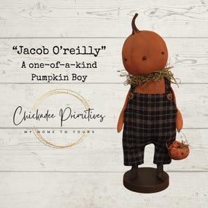 Op de afbeelding: Een handgemaakte pompoenfiguur genaamd "Jacob O'Reilly" met een pompoenhoofd, draagt een geruite overall en houdt een klein mandje vast. De figuur heeft een strooien kraag en staat op een houten voet. De tekst "Chickadee Primitives" is ook zichtbaar.