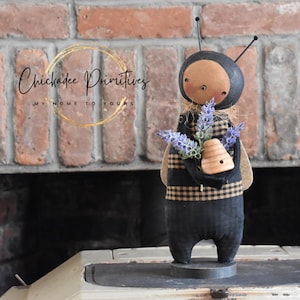 May include: A handcrafted bee doll with a black and tan checkered pattern on its body and wings. The doll holds a bouquet of purple flowers and a wooden beehive. The doll has a black head with antennae and is standing on a gray base.