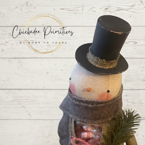 May include: A handmade snowman doll with a black top hat, a brown scarf, and a sprig of greenery in its hand. The snowman is holding a bundle of candy canes. The doll is standing on a white wood background. The text "Chickadee Primitives" is visible in the background.