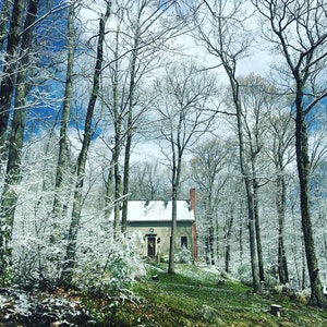 May include: A small house in a snowy forest setting. The house has a white roof, a red brick chimney, and a dark door. The trees are bare, with snow on the branches and ground. The sky is blue with some clouds, creating a serene winter scene.