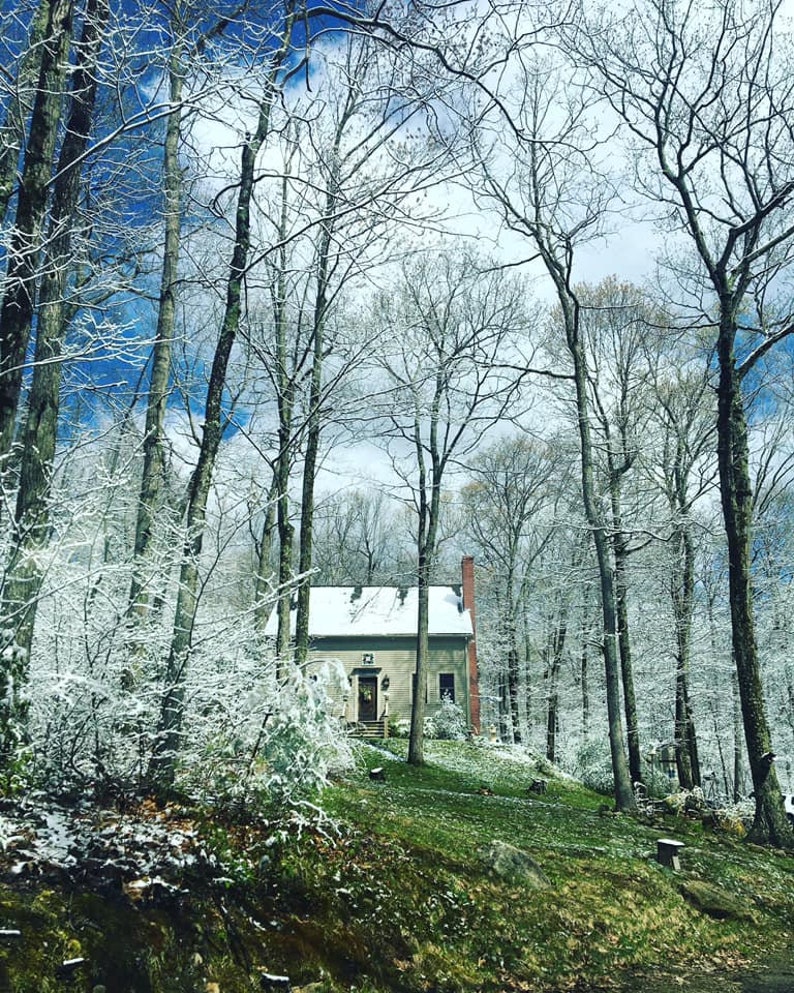 May include: A small house in a snowy forest setting. The house has a white roof, a red brick chimney, and a dark door. The trees are bare, with snow on the branches and ground. The sky is blue with some clouds, creating a serene winter scene.