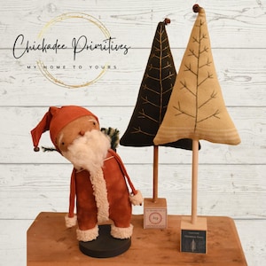 Pode incluir: Um boneco de Papai Noel feito à mão com um terno vermelho e barba branca fica ao lado de duas árvores de Natal de tecido. As árvores estão em bases de madeira com as palavras "Alegria" e "Fazenda de Árvores de Natal" impressas nelas. O fundo é um painel de madeira branqueada.