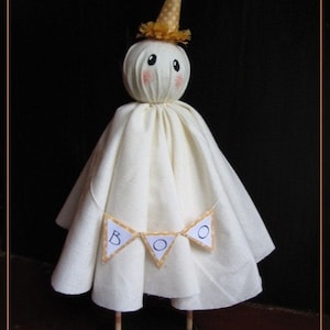 May include: A handmade Halloween ghost figure with a cream-colored body and a small, round head. The ghost wears a gold-colored party hat and a banner that spells "BOO". The figure is mounted on a black base with wooden dowel legs.