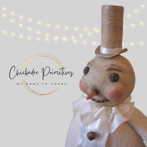 May include: A whimsical snowman figurine with a white top hat, a brown paper hat, and a white ruffle collar. The snowman has a painted face with rosy cheeks and a carrot nose. The figurine is made of fabric and has a rustic, handmade look.