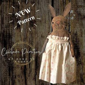 May include: A handmade fabric bunny doll wearing a white dress with pink polka dots. The doll has brown fabric for the body and face, with a stitched nose and black button eyes. The doll is standing against a wooden background with the text "NEW Pattern" and "Chickadee Primitives" visible.