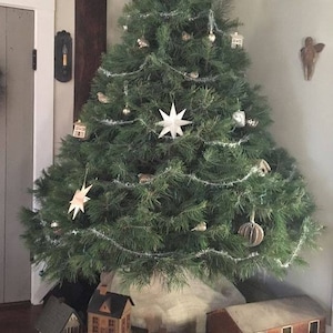 May include: A decorated Christmas tree with star and bird ornaments, tinsel, and paper star accents. Miniature wooden houses are arranged at the base of the tree. The tree is in a corner of a room with a neutral colour palette.