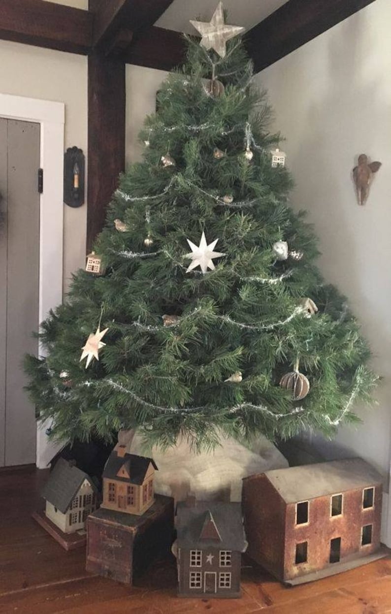 May include: A decorated Christmas tree with star and bird ornaments, tinsel, and paper star accents. Miniature wooden houses are arranged at the base of the tree. The tree is in a corner of a room with a neutral colour palette.