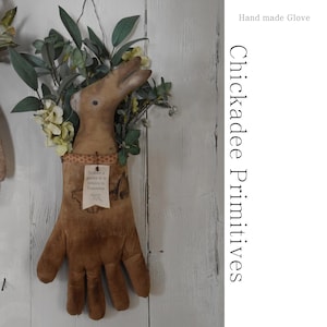 Könnte beinhalten: Eine braune Stoffhand mit einem kleinen Strauß weißer Blumen in der Handfläche. Die Hand hängt an einer weißen Holzwand. Der Text "To have a garden is to believe in tomorrow" ist auf einem Etikett gedruckt, das an der Hand befestigt ist. Der Text "Hand made Glove Chickadee Primitives" ist auf weißem Hintergrund gedruckt.