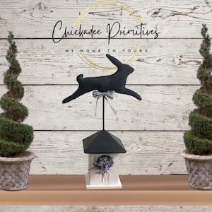 May include: Black bunny silhouette on a wooden stand with a birdhouse and two potted topiary trees. The sign above reads "Chickadee Primitives My Home to Yours".