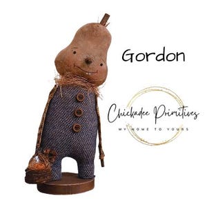 May include: A handcrafted doll named "Gordon" with a pear-shaped head and a stitched face. The doll wears a blue fabric outfit with three wooden buttons and a straw collar. It holds a small basket and stands on a wooden base.