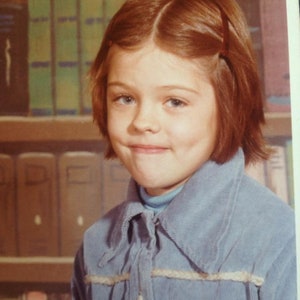 May include: A portrait of a young girl with auburn hair and blue eyes. She is wearing a light blue corduroy shirt over a light blue turtleneck. A bookshelf filled with books is in the background.
