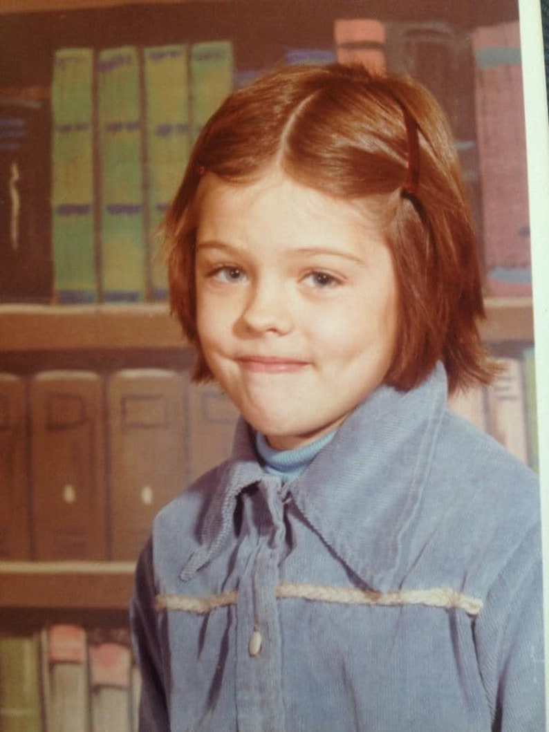 May include: A portrait of a young girl with auburn hair and blue eyes. She is wearing a light blue corduroy shirt over a light blue turtleneck. A bookshelf filled with books is in the background.