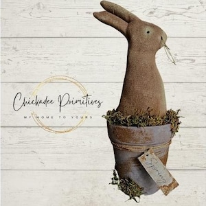 Op de afbeelding: Een bruin stoffen konijn zit in een kleine terracotta pot met mos en een touwaccent. De pot heeft een label met de tekst "Lente". De achtergrond is een witgeschilderd houten oppervlak met de tekst "Chickadee Primitives My Home To Yours" in zwart.