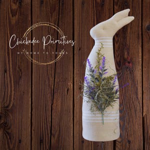 Peut inclure: Un lapin en tissu beige avec un motif de rayures blanches et un bouquet de fleurs violettes dans ses bras. Le lapin est debout sur un fond en bois brun. Le texte "Chickadee Primitives" est visible dans le coin supérieur gauche de l'image.
