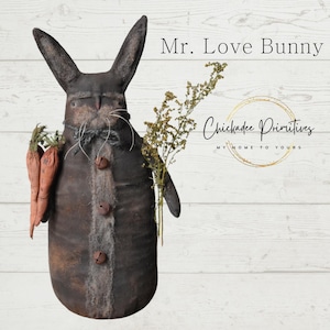 Könnte beinhalten: Ein braunes und graues Kaninchen mit langen Ohren, das zwei Karotten hält. Das Kaninchen trägt ein grau-weißes Hemd mit Knöpfen und Glöckchen. Das Kaninchen steht auf einem weißen Holzgrund. Der Text "Mr. Love Bunny" ist in Schwarz über dem Kaninchen geschrieben. Der Text "Chickadee Primitives" ist in Gold unter dem Kaninchen geschrieben. Der Text "My Home to Yours" ist in Schwarz unter dem goldenen Text geschrieben.