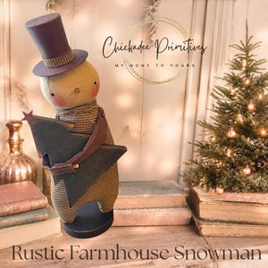 May include: A fabric snowman figurine wearing a dark top hat and a brown checkered body, holding a dark green wooden Christmas tree cutout. The snowman has pink cheeks and a small orange nose. Text reads Chickadee Primitives.