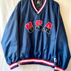 Peut inclure: Veste de survêtement Starter bleu marine avec col en V. Le mot "USA" est affiché sur la poitrine en lettres rouges, noires et blanches. La veste a des bordures rayées blanches et rouges sur les poignets et la taille.