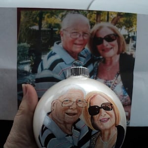 May include: A white glass ornament with a painted image of two older adults. The ornament features a man and a woman with gray hair and smiling. The man is wearing a blue and white striped shirt and the woman is wearing a white shirt with a black and white patterned design.