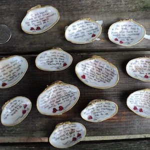 May include: Hand-painted seashells with gold trim and inspirational quotes. Each shell features handwritten text, red heart accents, and a unique design. The shells are arranged on a rustic wooden surface, with a coin for scale.