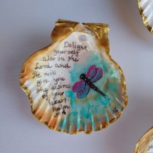 Peut inclure: Une coquille de coquillage avec une bordure dorée et une libellule peinte. La coquille porte une citation manuscrite : "Délectez-vous aussi dans le Seigneur, et il vous donnera les désirs de votre cœur. Psaume 31:4".