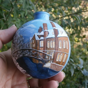 May include: A hand-painted Christmas ornament depicting a winter scene with a bridge, a building, and trees. The ornament is blue, white, and brown.