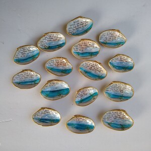 May include: A collection of painted seashells with gold trim. Each shell features a small landscape painting of a beach scene with blue water and a light blue sky. Handwritten Bible verses are included on each shell.