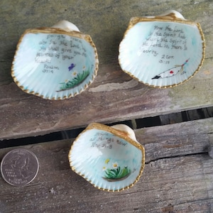 Może przedstawiać: Trzy ręcznie malowane muszle z pozłacanymi obramowaniami i inspirującymi cytatami. Muszle są pomalowane na jasnoniebiesko z kwiatowymi akcentami i tekstami z Biblii. Jedna muszla ma ilustrację motyla.