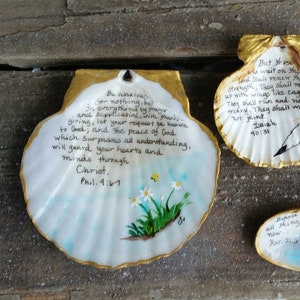 May include: Three painted seashells with gold trim. The largest shell has the text "Be anxious for nothing..." and a floral design. The other two shells have inspirational quotes and small paintings. A quarter is next to the largest shell.