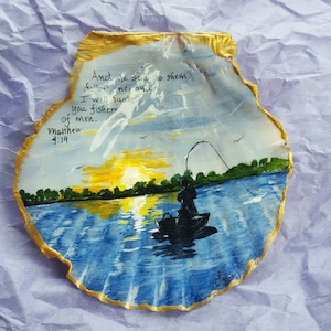 Könnte beinhalten: Eine Muschel, die mit einer Sonnenuntergangsszene eines Menschen beim Angeln auf einem See bemalt ist. Die Muschel ist mit Gold eingefasst und trägt den Text "And he said to them, follow me, and I will make you fishers of men. Matthew 4:19" auf.