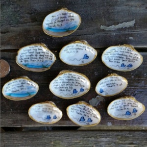 Może przedstawiać: Ręcznie malowane muszle z złotym obramowaniem, z inspirującymi cytatami i małymi niebieskimi motywami serca. Muszle mają namalowane sceny oceanu i nieba. Tekst zawiera wersety biblijne. Muszle są ułożone na drewnianej powierzchni.