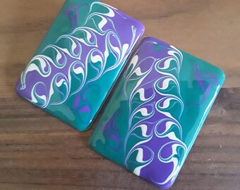 May include: Two rectangular coasters with a teal base and a swirling pattern in white, purple, and teal. The coasters have a glossy finish and are displayed on a wooden surface. The design is abstract and artistic.