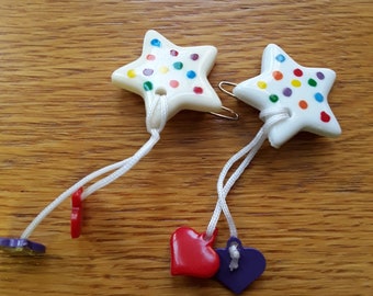 May include: Two white ceramic star-shaped ornaments with colorful polka dots and white string attached to each. One string has a red heart charm and the other has a purple heart charm.