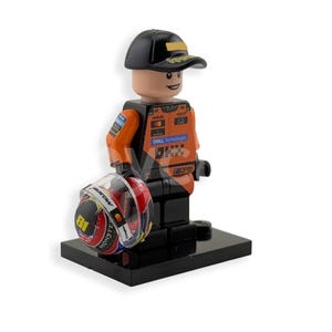 May include: A Lego figurine of a race car driver in a black cap with gold trim and an orange racing suit. The figurine holds a red, white, and black helmet with the number 81. The figurine is on a black base.
