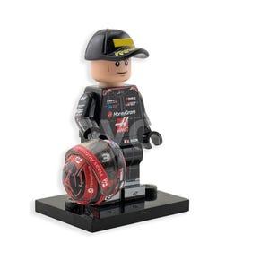 Op de afbeelding: Een Lego-figuur van een autocoureur met een zwarte pet met een gele streep en een zwart racepak. Het figuur houdt een rood-zwarte helm vast en staat op een zwarte basis.