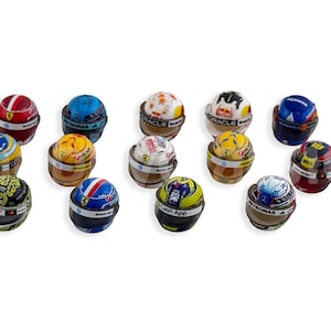 Peut inclure: Une collection de mini casques de course, chacun avec des designs uniques et des logos de sponsors. Les casques présentent une variété de couleurs, dont le rouge, le bleu, le jaune et le noir, sur un fond blanc. Ces objets de collection sont liés à la Formule 1.