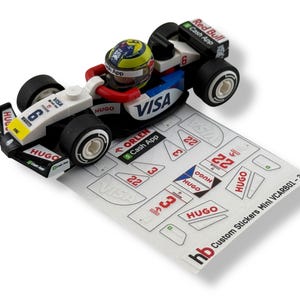 May include: A miniature Formula 1 race car model with a driver in a helmet. The car is white, black, and blue, with sponsor logos like VISA and Red Bull. A sheet of custom stickers is next to the car.