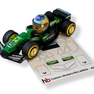 Könnte beinhalten: Ein grünes und schwarzes Spielzeug-Rennauto mit der Nummer 14 und Sponsorenlogos. Ein Miniaturfahrer mit Helm sitzt im Cockpit. Enthält einen Bogen mit individuellen Aufklebern. Der Text "Custom Stickers Mini AMR24 - 2024" ist sichtbar.
