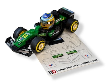 May include: A green and black toy race car with the number 14 and sponsor logos. A miniature driver wearing a helmet sits in the cockpit. Includes a sheet of custom stickers. The text "Custom Stickers Mini AMR24 - 2024" is visible.