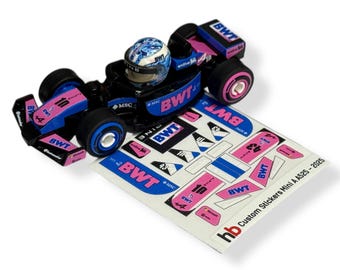 Peut inclure: Une voiture de course jouet noire et bleue avec des accents roses et un casque assorti. La voiture porte le numéro 10 et la marque BWT. Une feuille d'autocollants personnalisés est à côté de la voiture.