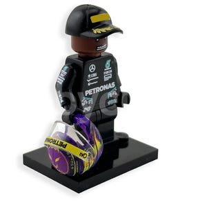 Może przedstawiać: Czarna figurka Lego przedstawiająca kierowcę Formuły 1 w czapce z żółtym paskiem. Figurka ma na sobie czarny kombinezon wyścigowy z logo Petronas i trzyma kask z fioletowo-żółtym wzorem.