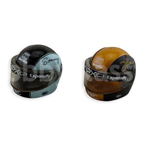 Puede incluir: Dos cascos de carreras en miniatura, uno negro y azul, el otro dorado y negro. Ambos tienen viseras y el texto "PXG Expensify" e "IWC". El casco negro y azul tiene el nombre "Sonny".