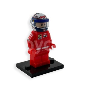 May include: A red Lego minifigure of a racing driver wearing a red, white and blue helmet with a clear visor. The figure is standing on a black base.