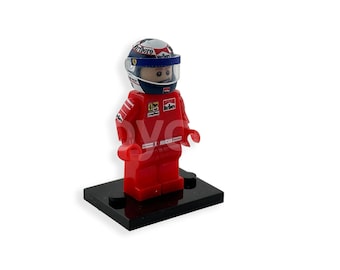 May include: A red Lego minifigure of a race car driver wearing a red, white, and blue helmet with a clear visor. The figure is standing on a black base.
