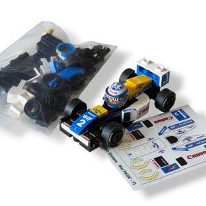 May include: A toy race car in white, blue, and yellow, with black wheels and a matching helmet. The car displays the number 2 and the word "Canon". A sheet of stickers is next to the car. A bag of loose parts is in the background.