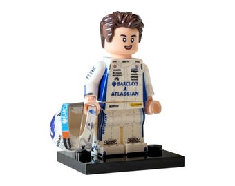 Peut inclure: Figurine Lego d'un pilote de course en uniforme blanc et bleu, tenant un casque. L'uniforme présente les logos "Barclays" et "Atlassian". La figurine est sur une base noire.