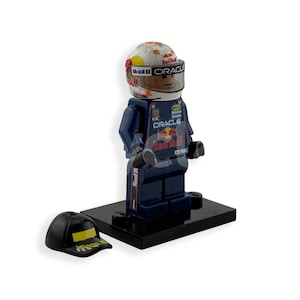 May include: A Lego figurine of a race car driver wearing a helmet with the Mobil 1 and Oracle logos. The figurine is in a dark blue racing suit with the Red Bull logo. A black baseball cap with a yellow design sits next to the figurine.